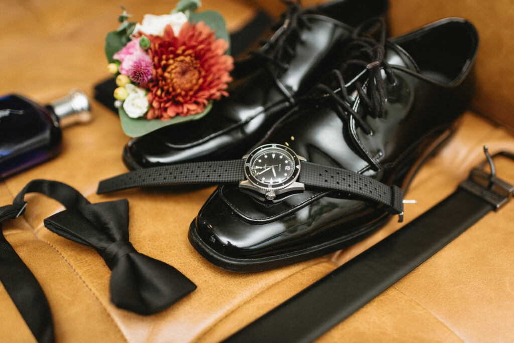 Grooms black wedding shoes with watch, belt, bowtie, polo blue cologne, and colorful pink, reds, and yellow boutonniere detail shot at The Farm Bakery and Events wedding venue in Quakertown, PA,