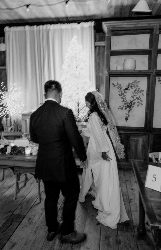 A candid, documentary style black and white image of a bride and groom walking toward their wedding cake at Terrain at Styers wedding venue. 