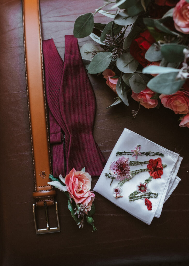 A grooms wedding details arrangement on an ottoman. A burgundy bowtie, light brown leather belt, wildflower floral pocket square, boutonniere, and a floral bouquet.