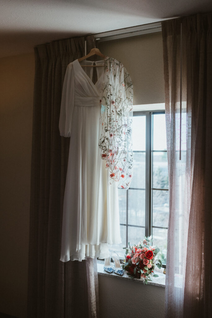 A brides wedding dress hanging from a hotel window with her wildflower laced floral veil hands with it. Arranged in the window sill are a bouquet and blue and white floral wedding heels. 