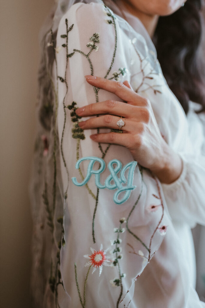 A brides custom wildflower laced floral veil that's monogrammed draped over her shoulder with her hand showing her engagement ring. 