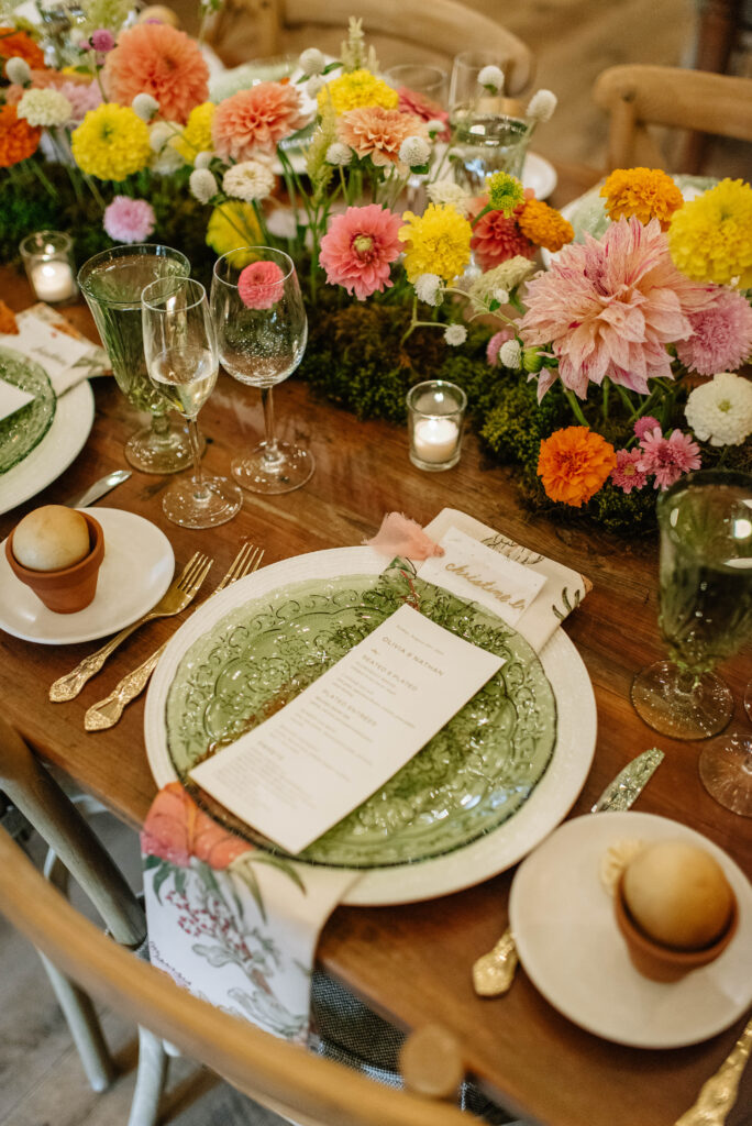 Stunning wedding reception details of colorful wildflower details, candles, green glass plates, wood chairs, ornate cups, and floral napkins at Terrain Gardens at Styers in Glen Mills, PA!